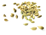 A scattered arrangement of green cardamom pods on a white surface. The pods vary in size and are randomly placed, creating a natural, organic pattern.