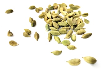 A scattered arrangement of green cardamom pods on a white surface. The pods vary in size and are randomly placed, creating a natural, organic pattern.