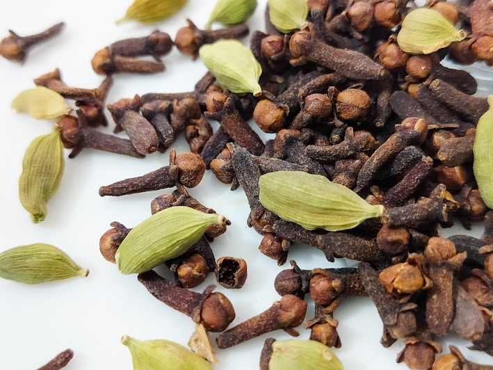 A close-up image of spices including cloves and cardamom pods scattered on a white surface. The cloves are dark brown with a unique elongated shape, while the cardamom pods are pale green and oval. The contrast between the light surface and the darker spices highlights their texture and color.