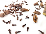 A collection of brown spices including cloves and cardamom pods scattered on a white surface. The cloves are small, dark, and have a distinctive shape with stems and heads. The cardamom pods are oblong with a rough texture, some of which are open to reveal small seeds inside. The arrangement of the spices is casual and spread across the image, creating an organic pattern.