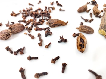 A collection of brown spices including cloves and cardamom pods scattered on a white surface. The cloves are small, dark, and have a distinctive shape with stems and heads. The cardamom pods are oblong with a rough texture, some of which are open to reveal small seeds inside. The arrangement of the spices is casual and spread across the image, creating an organic pattern.