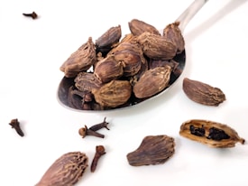 A metal spoon filled with dried cardamom pods is placed against a white background. Several individual pods and cloves are scattered around the spoon. The textures of the pods and cloves are rough and detailed, suggesting a natural, earthy theme.