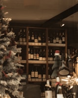Through a shop window, shelves are filled with a variety of wine bottles, categorized by country labels such as Italy and France. A decorated Christmas tree is visible on the left, adding a festive atmosphere. The lighting is warm and ambient.