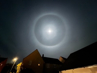 A bright circular halo surrounds the moon in a clear night sky, with residential houses in silhouette at the foreground. A street lamp emits a warm glow on the left, and foliage is partially illuminated by its light.