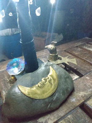 A decorative setup featuring a metallic crescent moon and an intricate bird figurine placed on a stone, with hanging crystal-like elements and dim lights in the background. The details are set on a wooden surface, creating an artistic and mystical atmosphere.