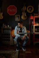 A person with curly hair sits on a couch holding an electric guitar in a dimly lit room. The walls feature a collection of music-related items, including guitars and posters. A 'Pare' sign, likely a stop sign, hangs on the wall, and there's a brightly colored Coca-Cola shelf nearby. The atmosphere is cozy and artistic.