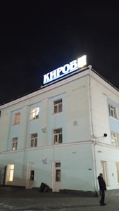 A light blue building with several windows is illuminated against the dark night sky. Atop the building, a bright sign with the word 'Киров' is prominently displayed. An individual stands near the entrance, and there are some items, possibly bags, placed beside the door.