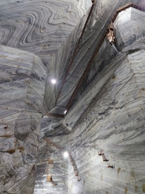 An expansive underground marble quarry features towering, textured marble walls with distinct swirling patterns and light fixtures mounted on them. Wooden ladders and platforms are scattered along the walls, indicating human activity and access.
