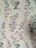A vintage floral wallpaper pattern featuring intricately designed flowers in shades of blue and pink intertwined with soft pink ribbons. The background is a light cream color that complements the gentle color palette of the florals. The design repeats vertically, creating a harmonious and continuous pattern across the surface.
