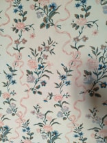 A vintage floral wallpaper pattern featuring intricately designed flowers in shades of blue and pink intertwined with soft pink ribbons. The background is a light cream color that complements the gentle color palette of the florals. The design repeats vertically, creating a harmonious and continuous pattern across the surface.