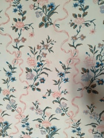 A vintage floral wallpaper pattern featuring intricately designed flowers in shades of blue and pink intertwined with soft pink ribbons. The background is a light cream color that complements the gentle color palette of the florals. The design repeats vertically, creating a harmonious and continuous pattern across the surface.