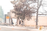 A demolished building with a partially collapsed structure is surrounded by debris and caution tape. A street sign warns of a stop, and barricades are placed on the road. Leafless trees and a lamp post stand nearby, under a hazy sky, indicating possible fire damage.