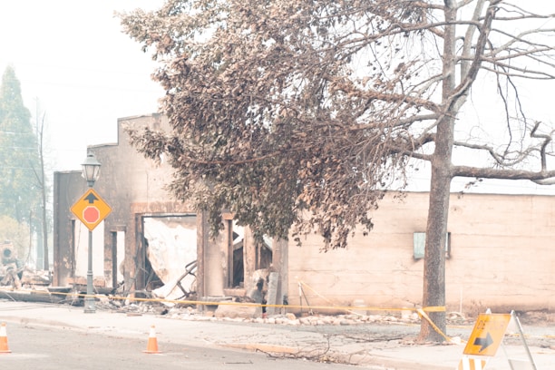 A demolished building with a partially collapsed structure is surrounded by debris and caution tape. A street sign warns of a stop, and barricades are placed on the road. Leafless trees and a lamp post stand nearby, under a hazy sky, indicating possible fire damage.