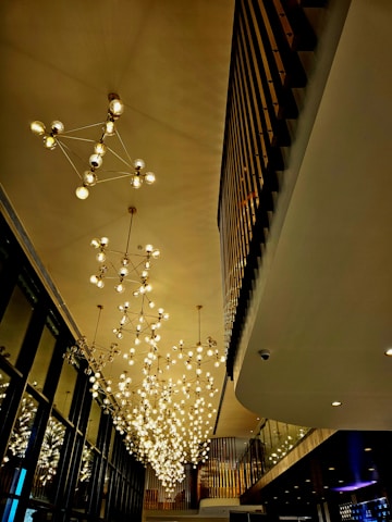 An elegant and spacious interior features a high ceiling adorned with modern, geometric chandeliers composed of multiple glowing bulbs. The design elements include a combination of linear and curvilinear structures with warm lights reflecting off glass and metal surfaces.