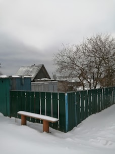 A snowy yard featuring a green metal fence and a wooden bench covered in snow. In the background, leafless trees and a rustic wooden shed with a sloping roof hint at a wintry rural setting, under an overcast sky.