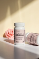 Two bottles of MultiFirst advanced formula adult multivitamins are displayed on a flat surface. The bottle in the foreground stands upright with legible text describing its benefits. The second bottle lies on its side, partially visible. A pink rose lies in the background, adding a soft touch to the composition. The lighting creates soft shadows and highlights on the surfaces.