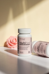 Two bottles of MultiFirst advanced formula adult multivitamins are displayed on a flat surface. The bottle in the foreground stands upright with legible text describing its benefits. The second bottle lies on its side, partially visible. A pink rose lies in the background, adding a soft touch to the composition. The lighting creates soft shadows and highlights on the surfaces.