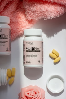 A bottle of Aforavo Essentials MultiFirst advanced formula adult multivitamin is centered in the image, surrounded by a pink fluffy fabric, yellow multi-vitamins, an open bottle spilling out more multi-vitamins, and a pink rose. The bottle label highlights various health benefits of the multivitamins.