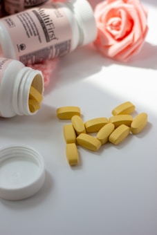 Several yellow tablets are scattered on a white surface next to an open white bottle, with another partially visible bottle in the background. A soft pink rose, possibly artificial, adds a delicate decorative touch.