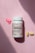 A bottle of advanced formula adult multivitamin labeled MultiFirst is placed on a soft pink surface. Next to the bottle are two yellow pills and three small, decorative pink flowers made of a material resembling resin or candy.