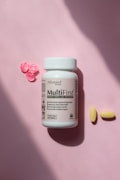 A bottle of advanced formula adult multivitamin labeled MultiFirst is placed on a soft pink surface. Next to the bottle are two yellow pills and three small, decorative pink flowers made of a material resembling resin or candy.