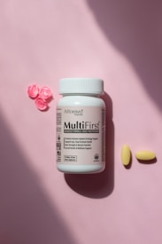 A bottle of advanced formula adult multivitamin labeled MultiFirst is placed on a soft pink surface. Next to the bottle are two yellow pills and three small, decorative pink flowers made of a material resembling resin or candy.