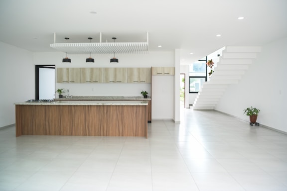 A spacious, modern kitchen with a large island featuring a marble countertop and wooden base. Sleek, minimalistic cabinetry in light wood tones lines the back wall. Black pendant lights hang from the ceiling above the island. The open layout extends to a bright area with white tile flooring and a geometric staircase on the right. Two potted plants add a touch of greenery to the minimalist decor.