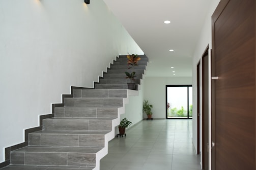 A modern interior hallway featuring a sleek gray tiled floor and a contemporary floating staircase. Several potted plants add a touch of greenery along the wall, and natural light streams in from a large window at the end of the hallway. Minimalist design with clean lines creates an open and airy feel.