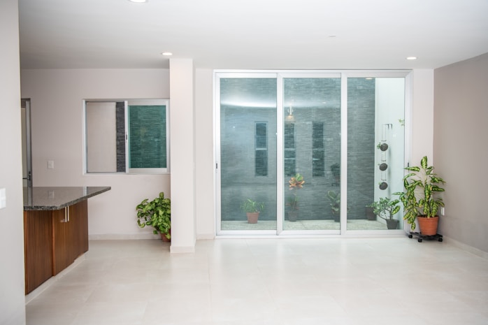 A modern, bright room with large sliding glass doors opening into a small garden area. Several potted plants, including leafy green varieties, are situated inside and outside the glass doors. The space has a sleek, minimalistic design with light-colored walls and flooring, as well as a small granite counter in the corner.