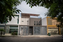 A modern urban house exterior with a clean and minimalistic design. The facade features geometric patterns with a combination of white and gray colors. Large windows and a metal gate add to the contemporary aesthetic. Surrounding the house are green leafy trees, providing a natural contrast.