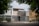 A modern urban house exterior with a clean and minimalistic design. The facade features geometric patterns with a combination of white and gray colors. Large windows and a metal gate add to the contemporary aesthetic. Surrounding the house are green leafy trees, providing a natural contrast.