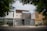 A modern urban house exterior with a clean and minimalistic design. The facade features geometric patterns with a combination of white and gray colors. Large windows and a metal gate add to the contemporary aesthetic. Surrounding the house are green leafy trees, providing a natural contrast.
