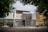 A modern urban house exterior with a clean and minimalistic design. The facade features geometric patterns with a combination of white and gray colors. Large windows and a metal gate add to the contemporary aesthetic. Surrounding the house are green leafy trees, providing a natural contrast.