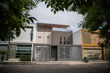 A modern urban house exterior with a clean and minimalistic design. The facade features geometric patterns with a combination of white and gray colors. Large windows and a metal gate add to the contemporary aesthetic. Surrounding the house are green leafy trees, providing a natural contrast.