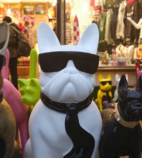 A white statue of a bulldog wearing black sunglasses and a black tie is prominently displayed in a shop or market with various colorful objects and apparel in the background. Other colorful bulldog statues surround the main figure, and a person is seen shopping in the background.