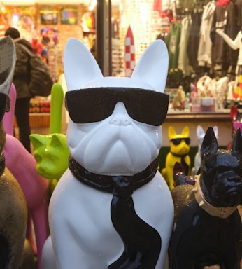 A white statue of a bulldog wearing black sunglasses and a black tie is prominently displayed in a shop or market with various colorful objects and apparel in the background. Other colorful bulldog statues surround the main figure, and a person is seen shopping in the background.