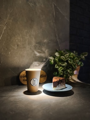 A cozy coffee cup with steam rising, placed on a wooden table next to a slice of carrot cake.
