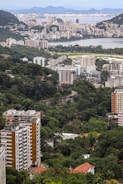 A sprawling urban landscape with a mixture of densely packed residential buildings and lush greenery. The city sprawls across the foreground with high-rise apartments and office buildings interspersed with parks and wooded areas. In the background, a body of water is visible, beyond which a distant cityscape merges with mountainous terrain under a partly cloudy sky.