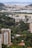 A sprawling urban landscape with a mixture of densely packed residential buildings and lush greenery. The city sprawls across the foreground with high-rise apartments and office buildings interspersed with parks and wooded areas. In the background, a body of water is visible, beyond which a distant cityscape merges with mountainous terrain under a partly cloudy sky.