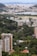 A sprawling urban landscape with a mixture of densely packed residential buildings and lush greenery. The city sprawls across the foreground with high-rise apartments and office buildings interspersed with parks and wooded areas. In the background, a body of water is visible, beyond which a distant cityscape merges with mountainous terrain under a partly cloudy sky.
