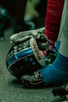 Close-up of a football helmet with a design featuring stars and stripes, a gloved hand resting on it, and a foot in a cleat nearby. The environment has a dim, greenish tint with a focus on the textures and materials.