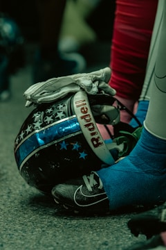 Close-up of a football helmet with a design featuring stars and stripes, a gloved hand resting on it, and a foot in a cleat nearby. The environment has a dim, greenish tint with a focus on the textures and materials.