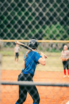 A person is standing at a baseball or softball field, ready to bat. The player is wearing a blue jersey, black pants, and a helmet. The scene is viewed through a fence, and there are other players visible in the background.