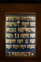 A display case contains an organized arrangement of various medals and decorations, each with a unique design, ribbon, and emblem. The medals are arranged in rows inside a wooden frame with a glass cover.