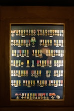 A display case contains an organized arrangement of various medals and decorations, each with a unique design, ribbon, and emblem. The medals are arranged in rows inside a wooden frame with a glass cover.