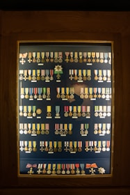 A display case contains an organized arrangement of various medals and decorations, each with a unique design, ribbon, and emblem. The medals are arranged in rows inside a wooden frame with a glass cover.