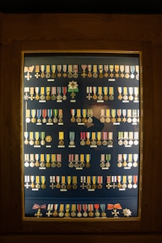 A display case contains an organized arrangement of various medals and decorations, each with a unique design, ribbon, and emblem. The medals are arranged in rows inside a wooden frame with a glass cover.