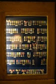 A wooden display case contains rows of various military medals and ribbons organized neatly. The medals are of different designs and colors, primarily gold and silver, and some feature stars or crosses. Many have colored ribbons attached, displaying shades of red, blue, yellow, and green. The background is a dark blue, enhancing the medals' visibility.