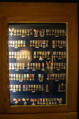 A wooden display case contains rows of various military medals and ribbons organized neatly. The medals are of different designs and colors, primarily gold and silver, and some feature stars or crosses. Many have colored ribbons attached, displaying shades of red, blue, yellow, and green. The background is a dark blue, enhancing the medals' visibility.