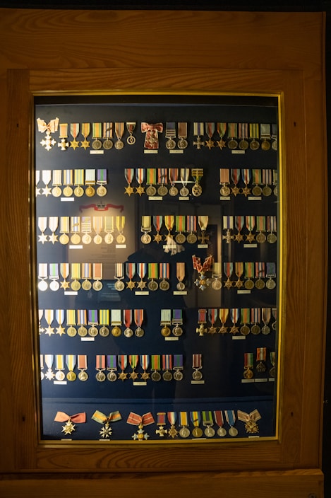 A wooden display case contains rows of various military medals and ribbons organized neatly. The medals are of different designs and colors, primarily gold and silver, and some feature stars or crosses. Many have colored ribbons attached, displaying shades of red, blue, yellow, and green. The background is a dark blue, enhancing the medals' visibility.